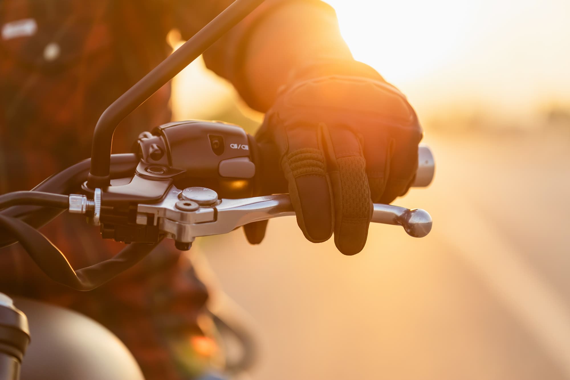 Gros plan sur le guidon d'une moto tenu par un motard équipé de gants de protection homologués avec un couché de soleil orangé en arrière plan