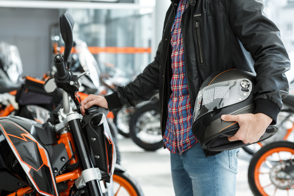 Jeune homme chez un concessionnaire de moto portant un casque homologué neuf sous le bras gauche et touchant l'avant d'une moto à vendre de la main droite